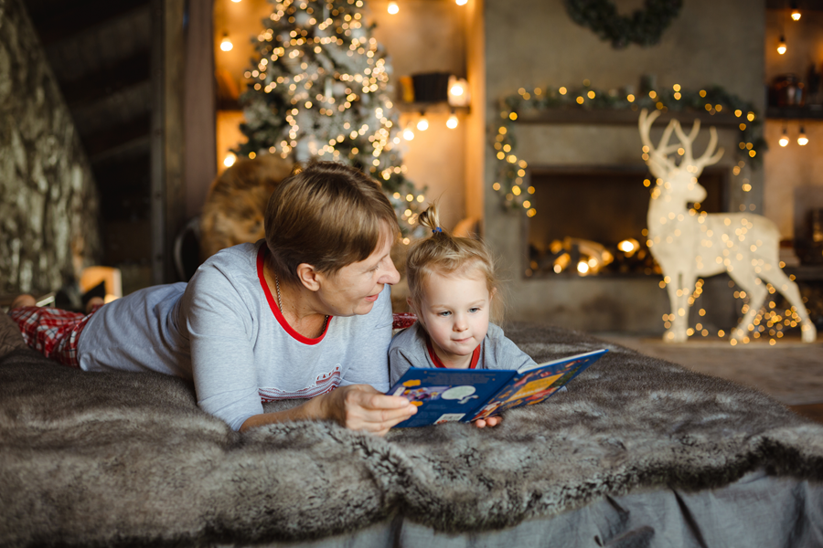 Grandparent reading a Christmas themed book to a child. They are wearing pyjamas and in the background are decorations.
